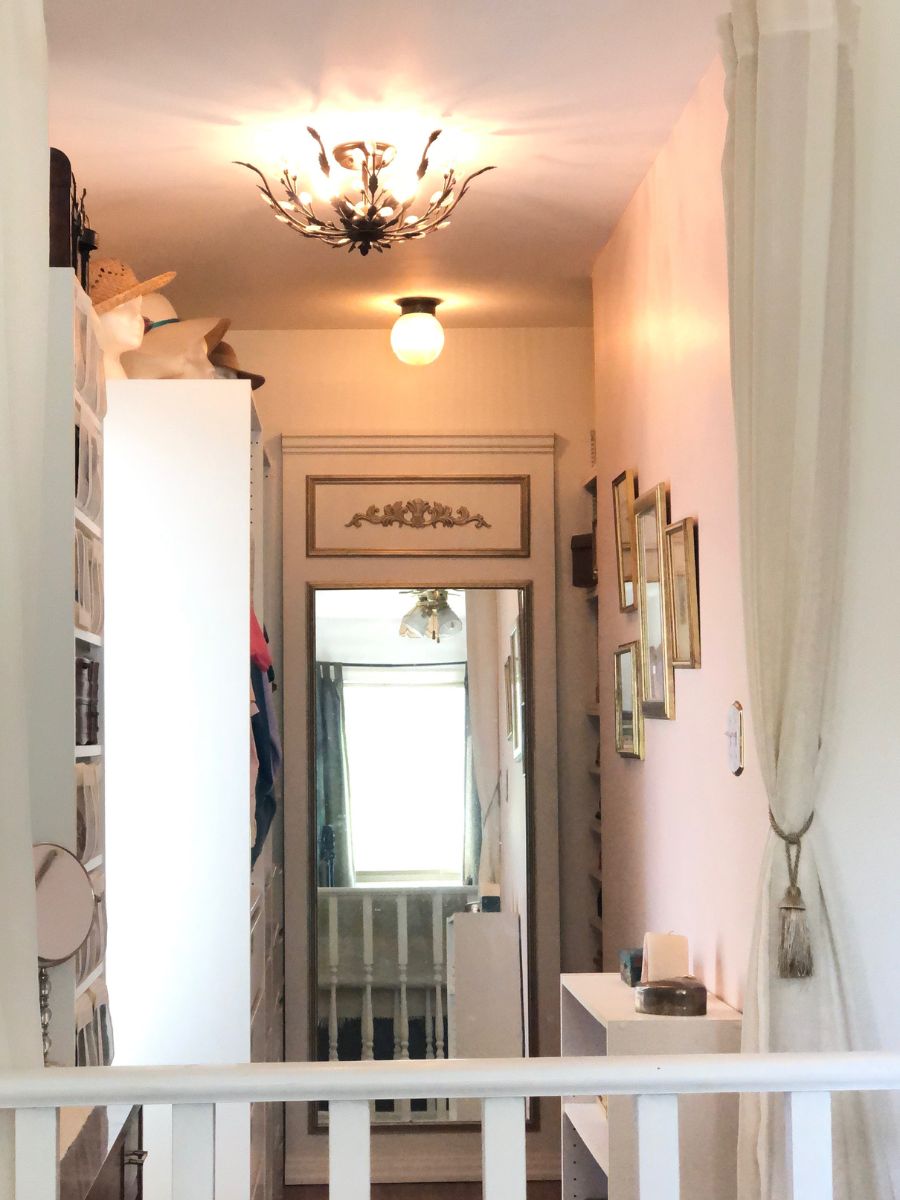 A photo of a dressing room created from a 5 foot wide dead end hallway. Open wardrobe units line the left side. Sunlight plus through from a window between two of them. On the right is a floor to ceiling alcove at the back slotted with shelves. Items in gold frames hang on the wall and there is a small bookcase in the foreground. The focal point of the photo and the closet is a large Trumeau Mirror in gold and white on the back wall.